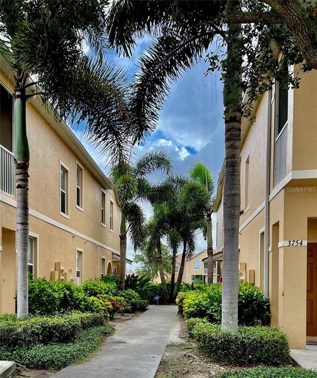 A concrete walkway leads between two tan residential buildings lined with palm trees — 3762 PARKRIDGE CIRCLE UNIT 23-106, SARASOTA, FL