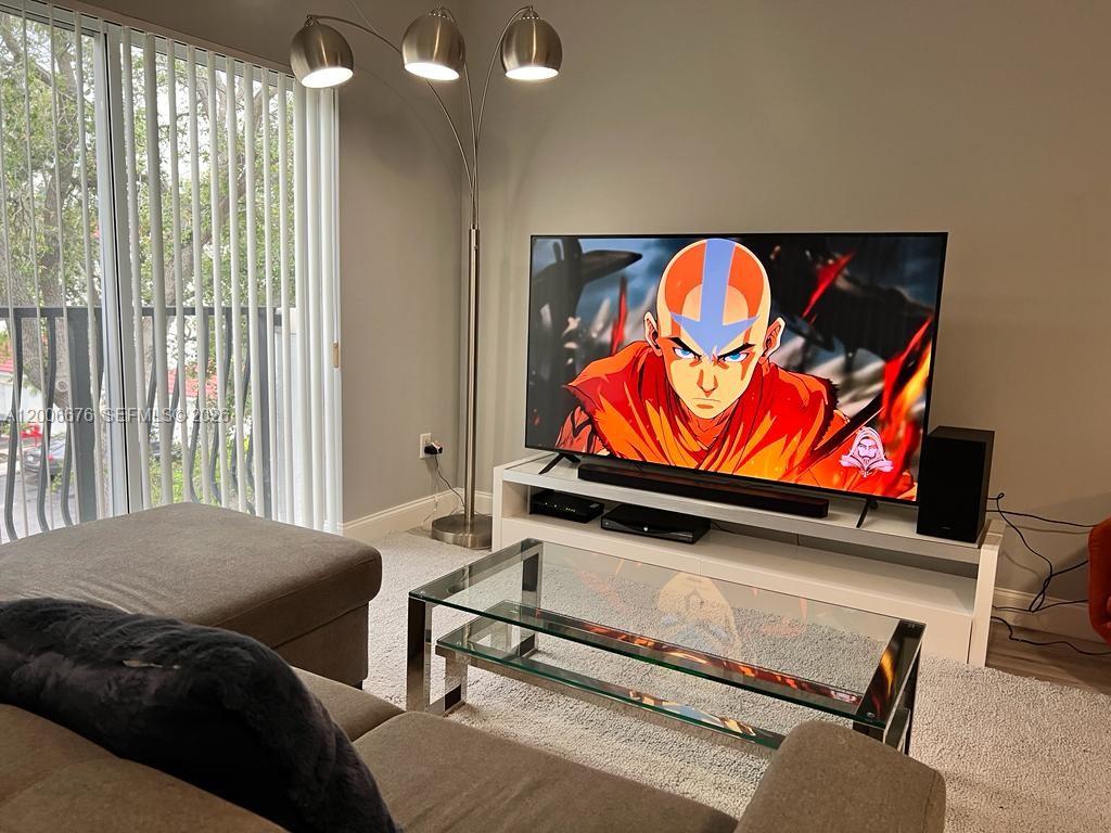 A living room features a television displaying an animated character, a glass coffee table, and lamps — AVENTURA, FL