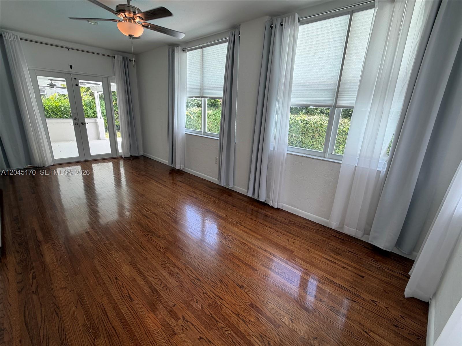 An empty room features polished hardwood floors, a ceiling fan, and large windows with curtains — 1420 Baracoa Ave, Coral Gables FL 33146, CORAL GABLES, FL