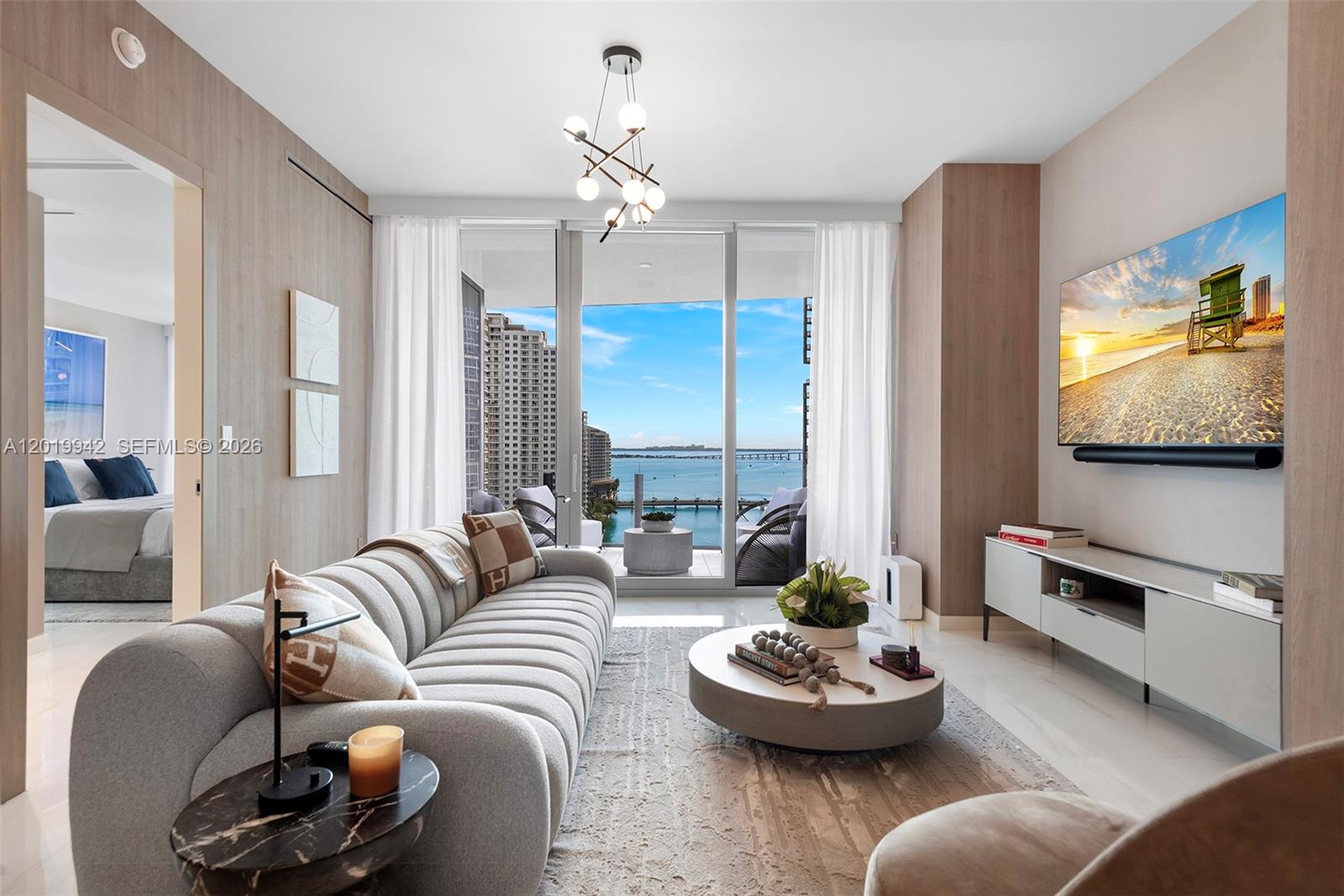 A modern living room features a curved sofa, circular coffee table, and balcony with water views — 300 Biscayne Blvd Way # 1605, Miami FL 33131, MIAMI, FL