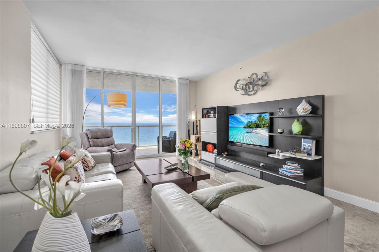 A living room features white leather sofas, a dark entertainment center, and a balcony view — 704 N Ocean Blvd # 804, Pompano Beach FL 33062, POMPANO BEACH, FL