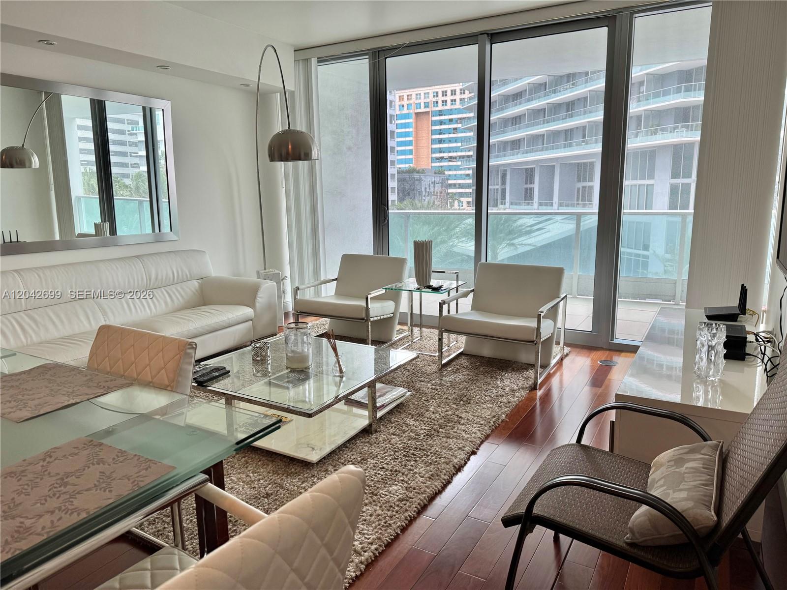 A modern living room features white leather furniture, a glass coffee table, and floor-to-ceiling windows — 1331 Brickell Bay Dr # 406, Miami FL 33131, MIAMI, FL