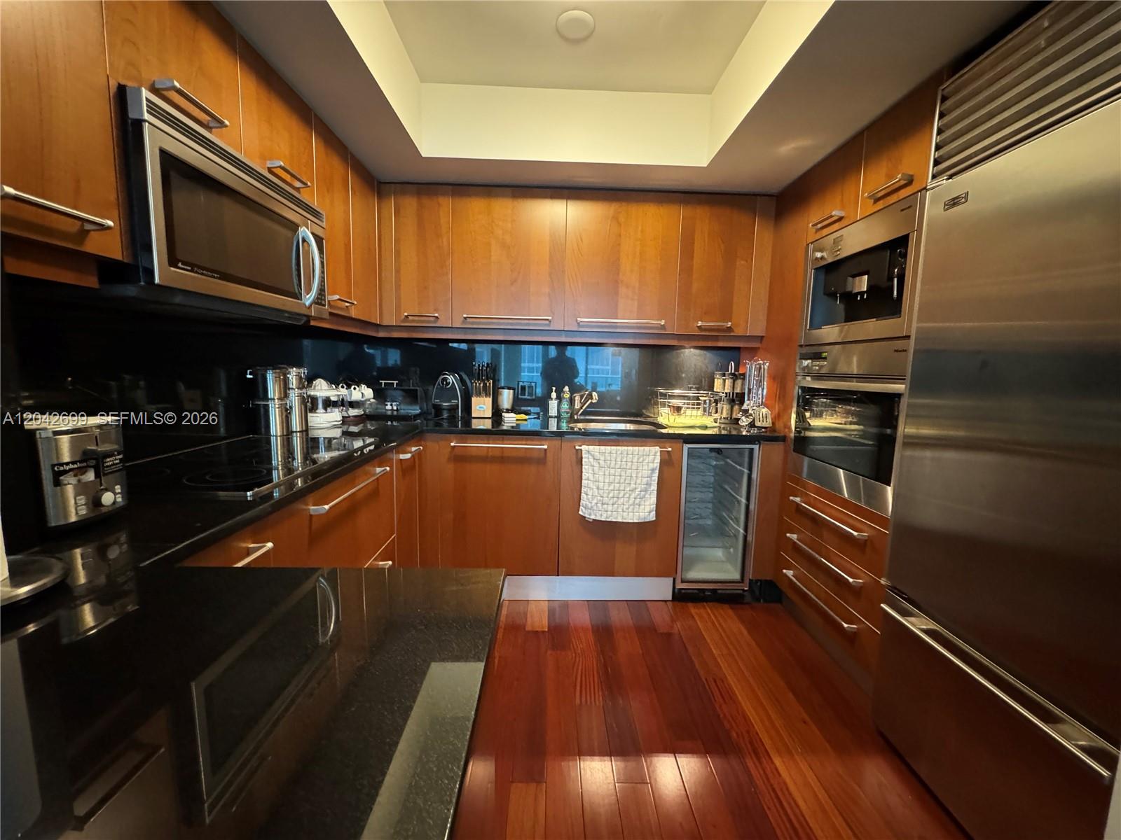 A kitchen features wood cabinetry, black countertops, stainless steel appliances, and hardwood flooring — 1331 Brickell Bay Dr # 406, Miami FL 33131, MIAMI, FL