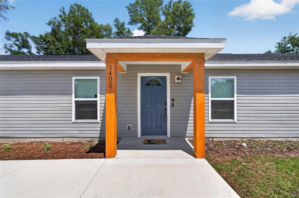 A gray house exterior features a blue front door under a wooden-pillared porch — 14027 SE 43RD TERRACE, SUMMERFIELD, FL