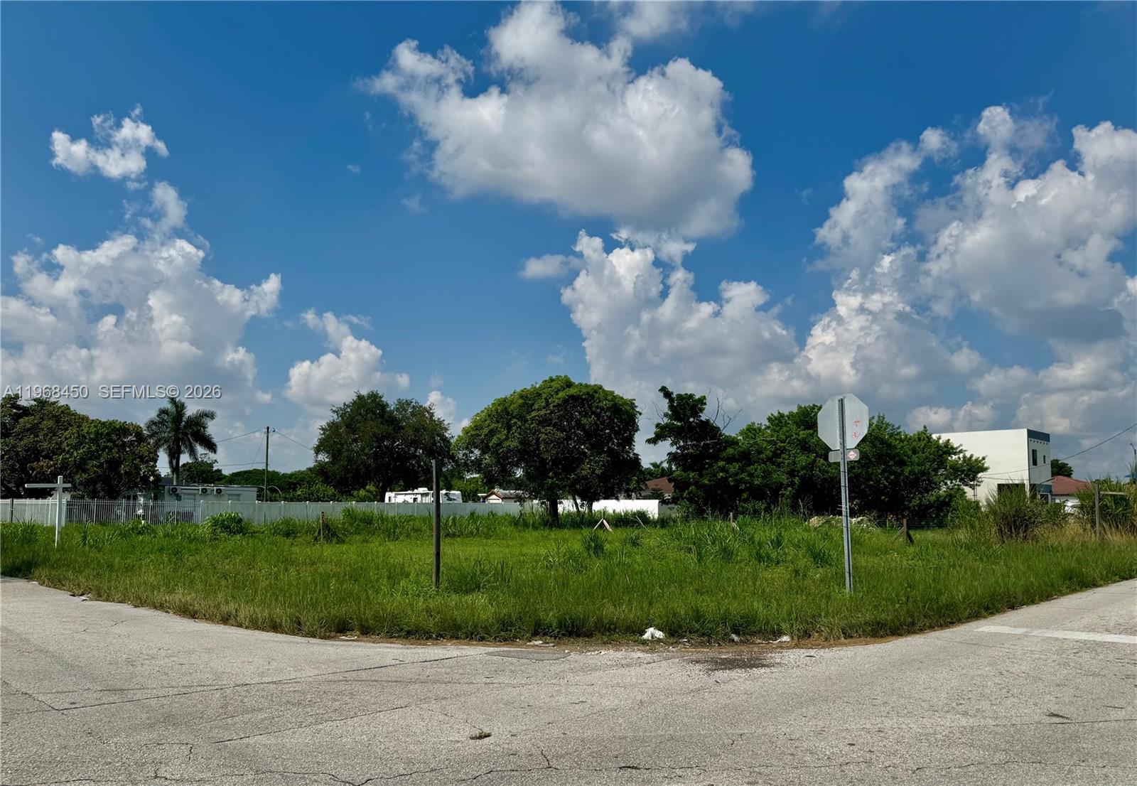 A vacant, grassy corner lot is bordered by paved roads and mature trees under a sky — 12208 SW 220 ST, Miami FL 33170, MIAMI, FL