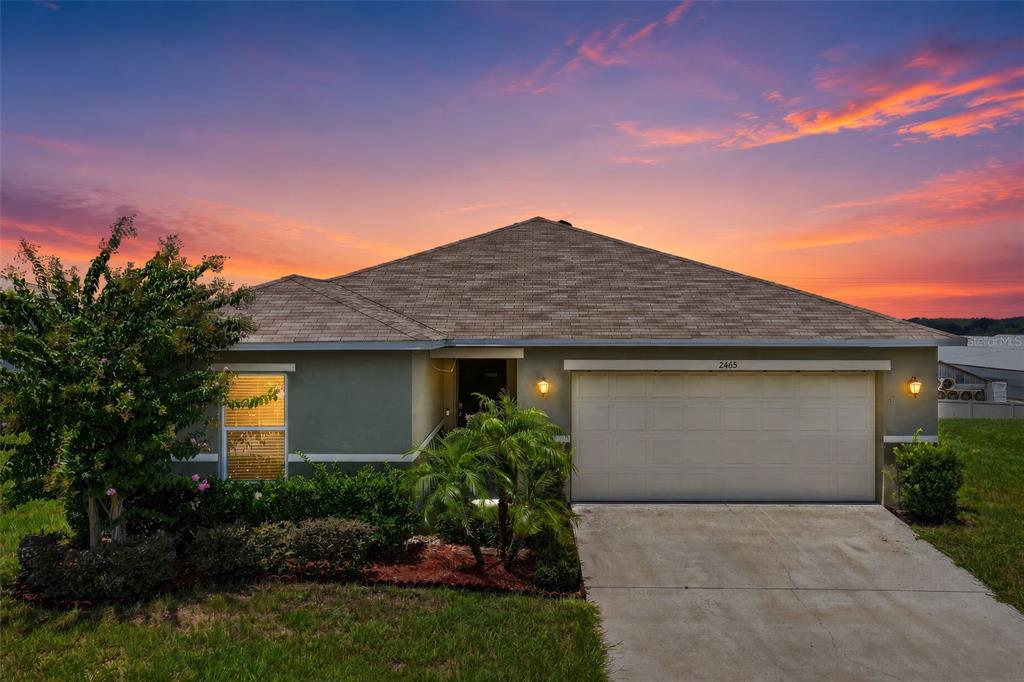 A single-story house with a two-car garage and a landscaped front yard at sunset — 2465 BEGONIA STREET, MASCOTTE, FL