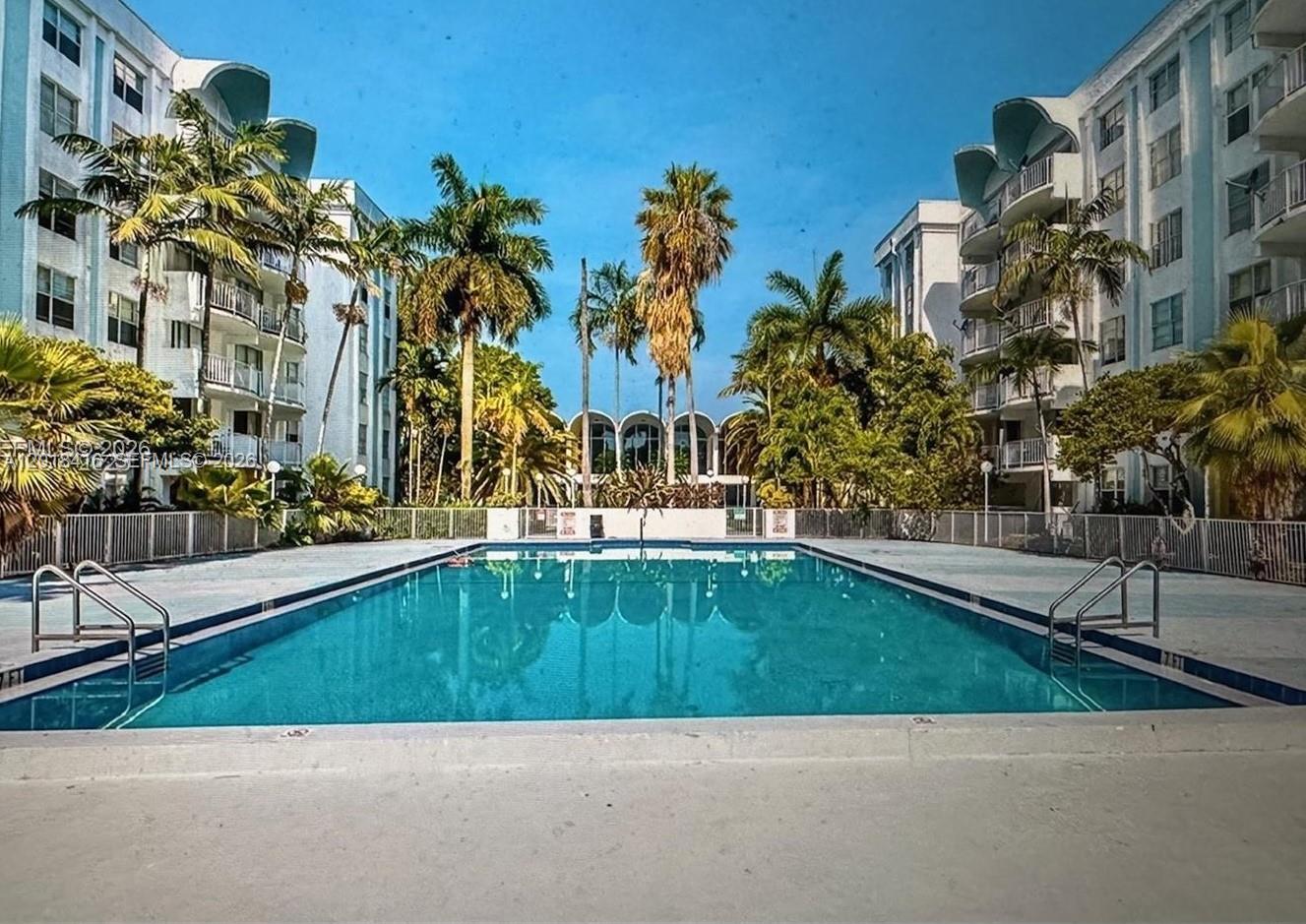 A rectangular swimming pool sits between two multi-story apartment buildings surrounded by palm trees — 496 NW 165th St Rd # 410, Miami FL 33169, MIAMI, FL