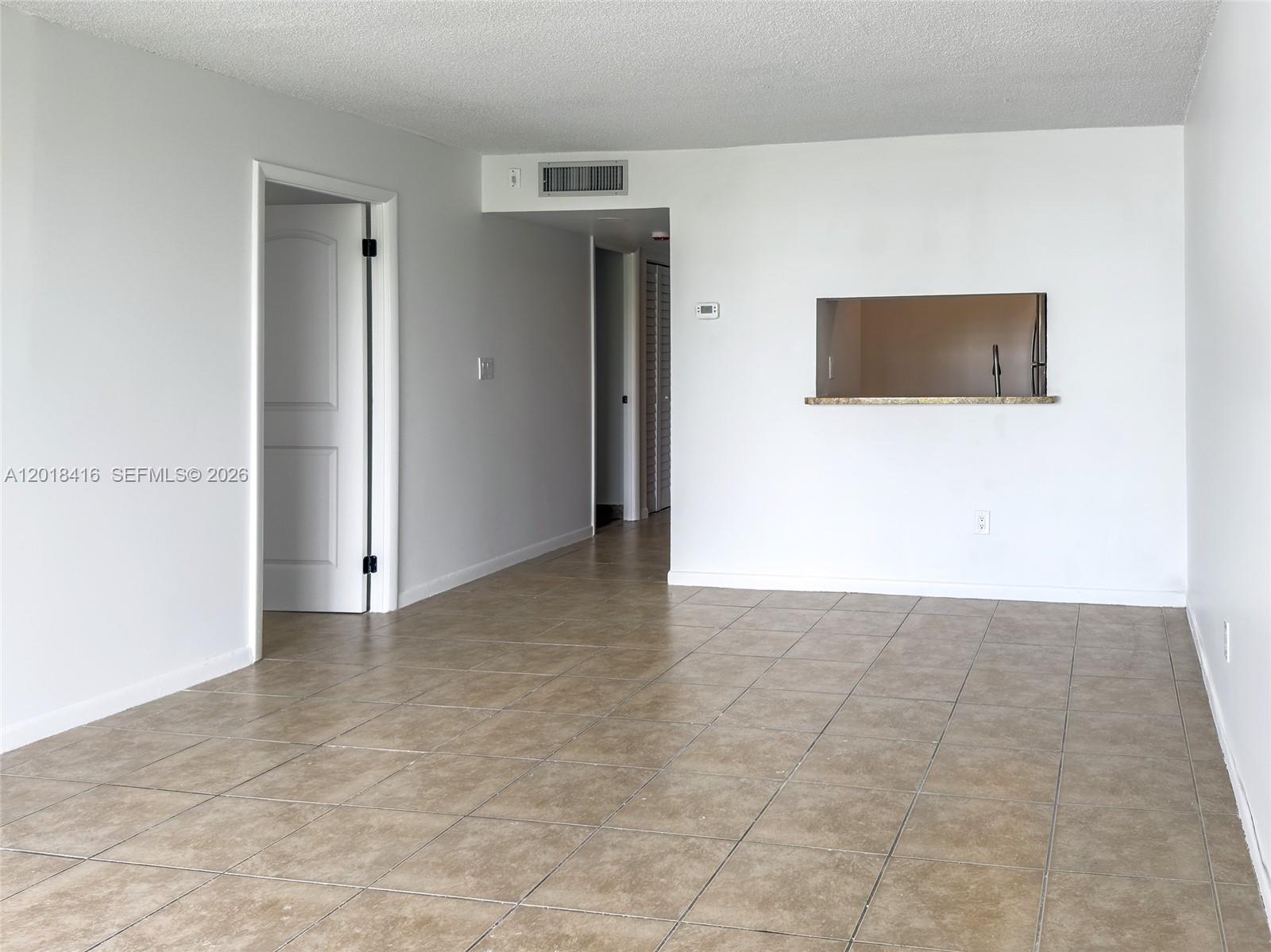 An empty room features beige tile flooring, white walls, and a kitchen pass-through window — 496 NW 165th St Rd # 410, Miami FL 33169, MIAMI, FL