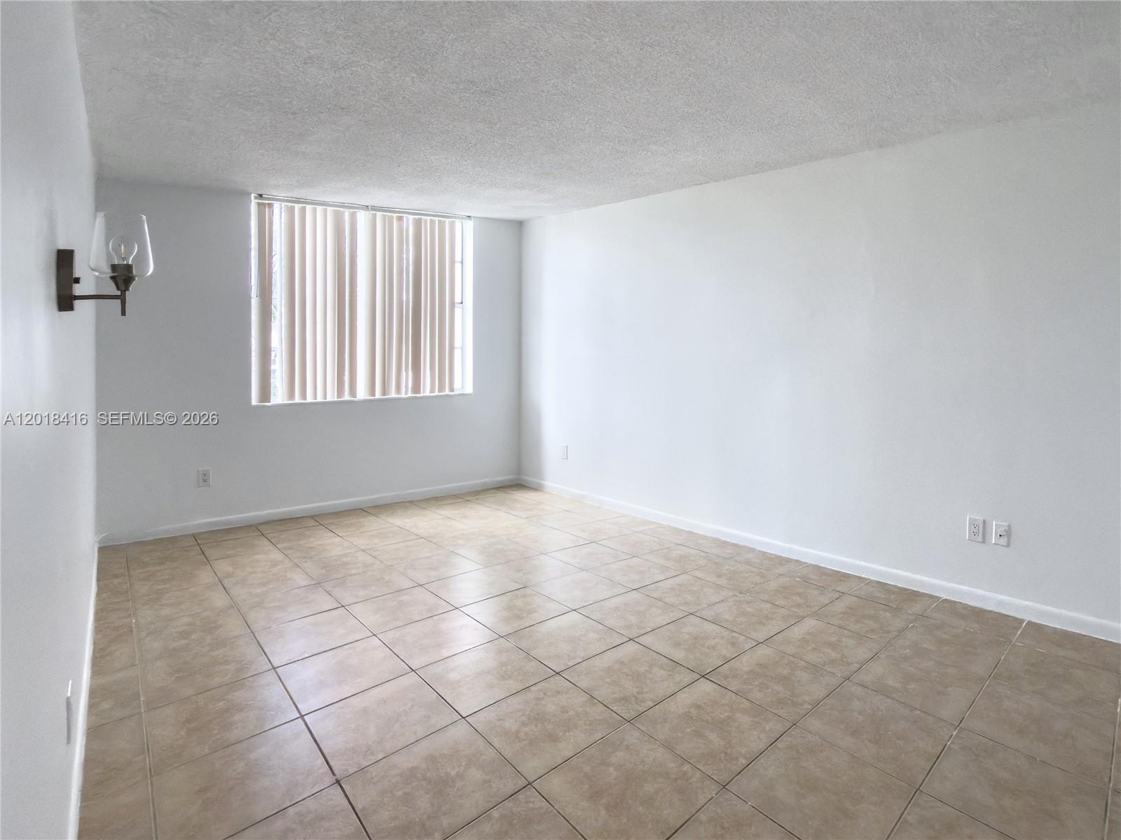 An empty room features beige tile flooring, white walls, a textured ceiling, and a window — 496 NW 165th St Rd # 410, Miami FL 33169, MIAMI, FL