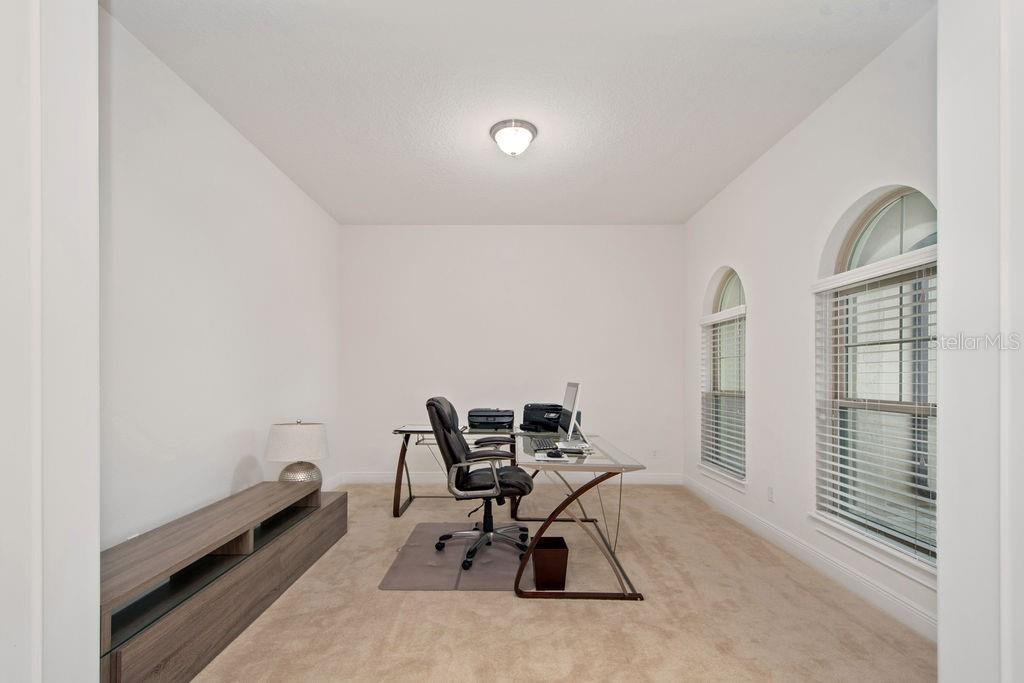 A home office features beige carpeting, white walls, a glass desk, and two arched windows — 17538 BLACK RAIL STREET, WINDERMERE, FL