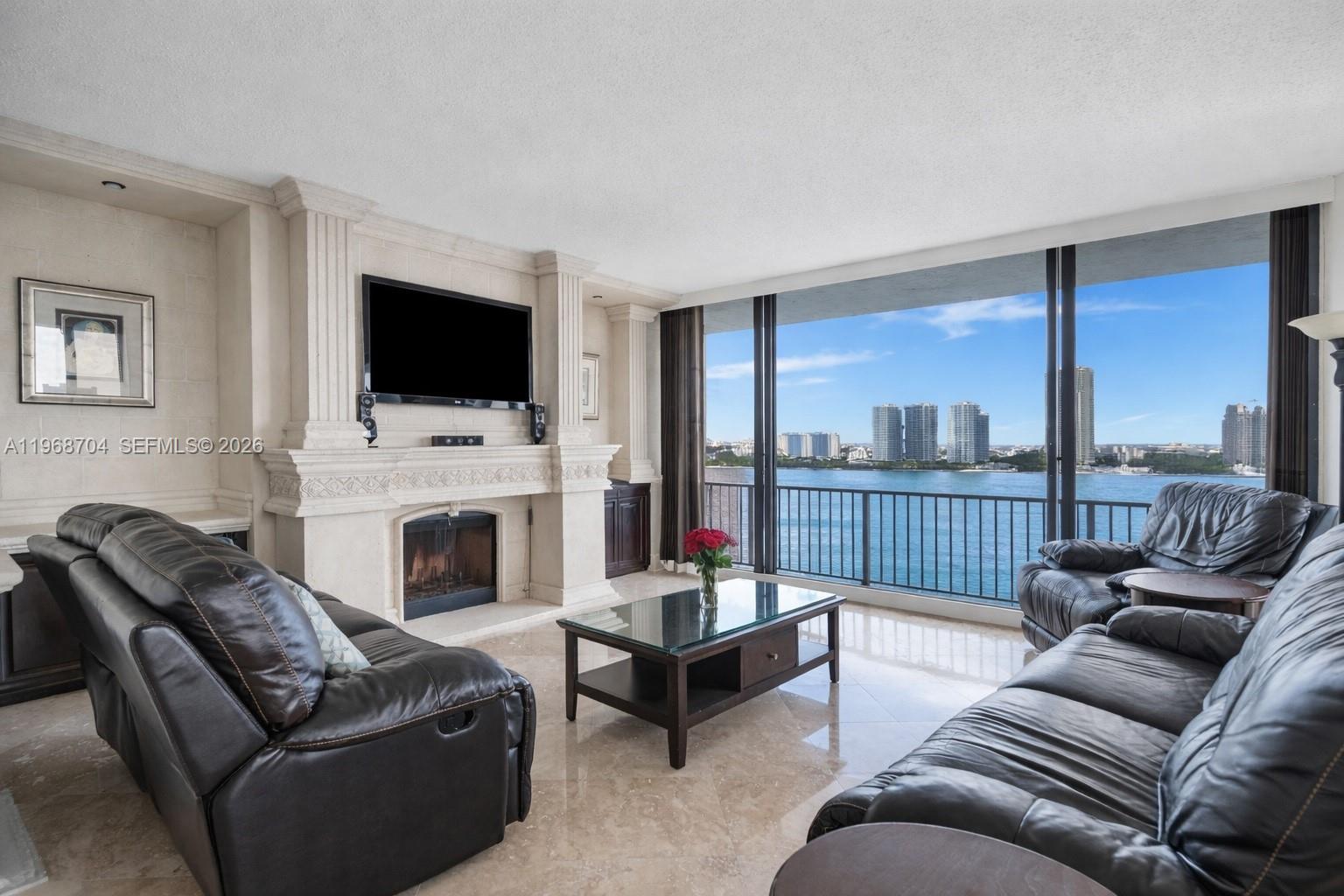 A living room features leather sofas, a fireplace, and floor-to-ceiling windows overlooking the water — 300 Three Islands Blvd, Hallandale Beach FL 33009, HALLANDALE BEACH, FL