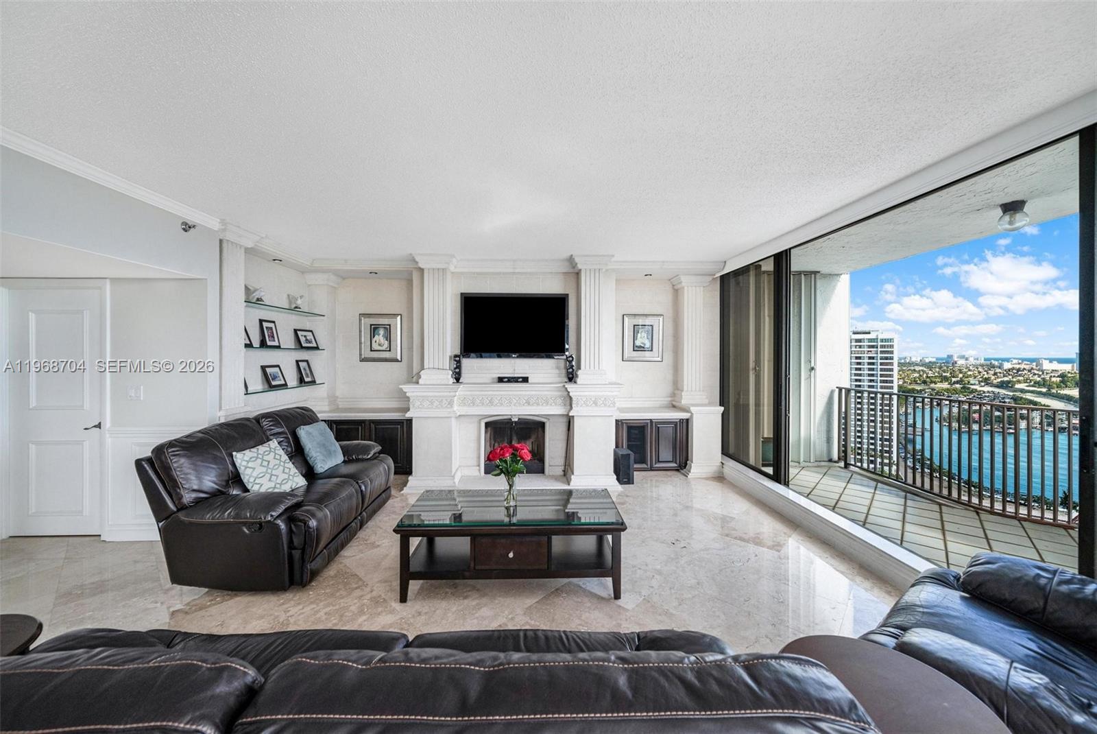 A living room features a fireplace, built-in shelving, leather sofas, and a balcony with water views — 300 Three Islands Blvd, Hallandale Beach FL 33009, HALLANDALE BEACH, FL