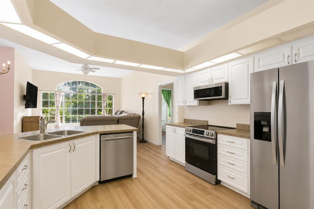 A kitchen with white cabinets, stainless steel appliances, and wood-look flooring opens to a living area — 12213 ROSELAND DRIVE, NEW PORT RICHEY, FL