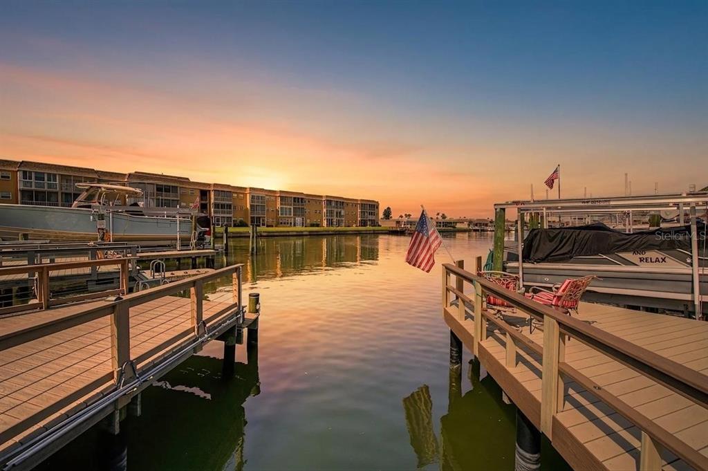 A view of a canal at sunset with wooden docks, boats, and a multi-story apartment building — 144 BOCA CIEGA POINT BOULEVARD N, ST PETERSBURG, FL