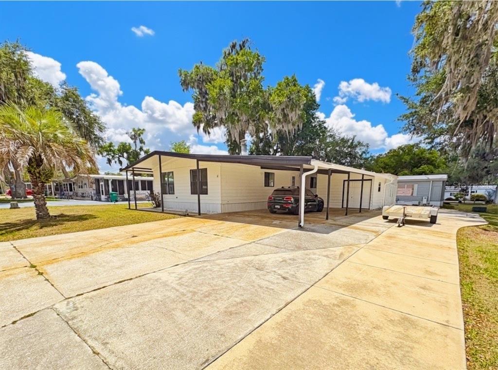 A single-story mobile home with a large concrete driveway, carport, and surrounding trees — 4 E QUAIL RUN, WILDWOOD, FL