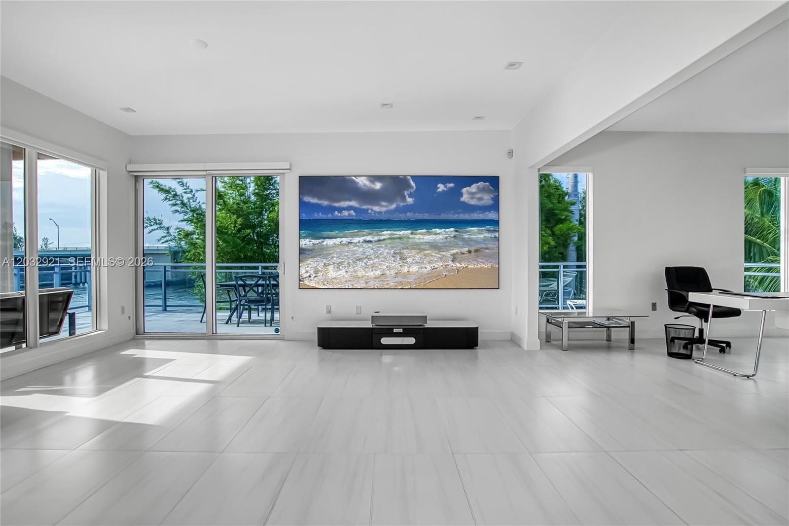 A bright, minimalist living room features white tile floors, a large wall-mounted screen, and balcony access — HOLLYWOOD, FL