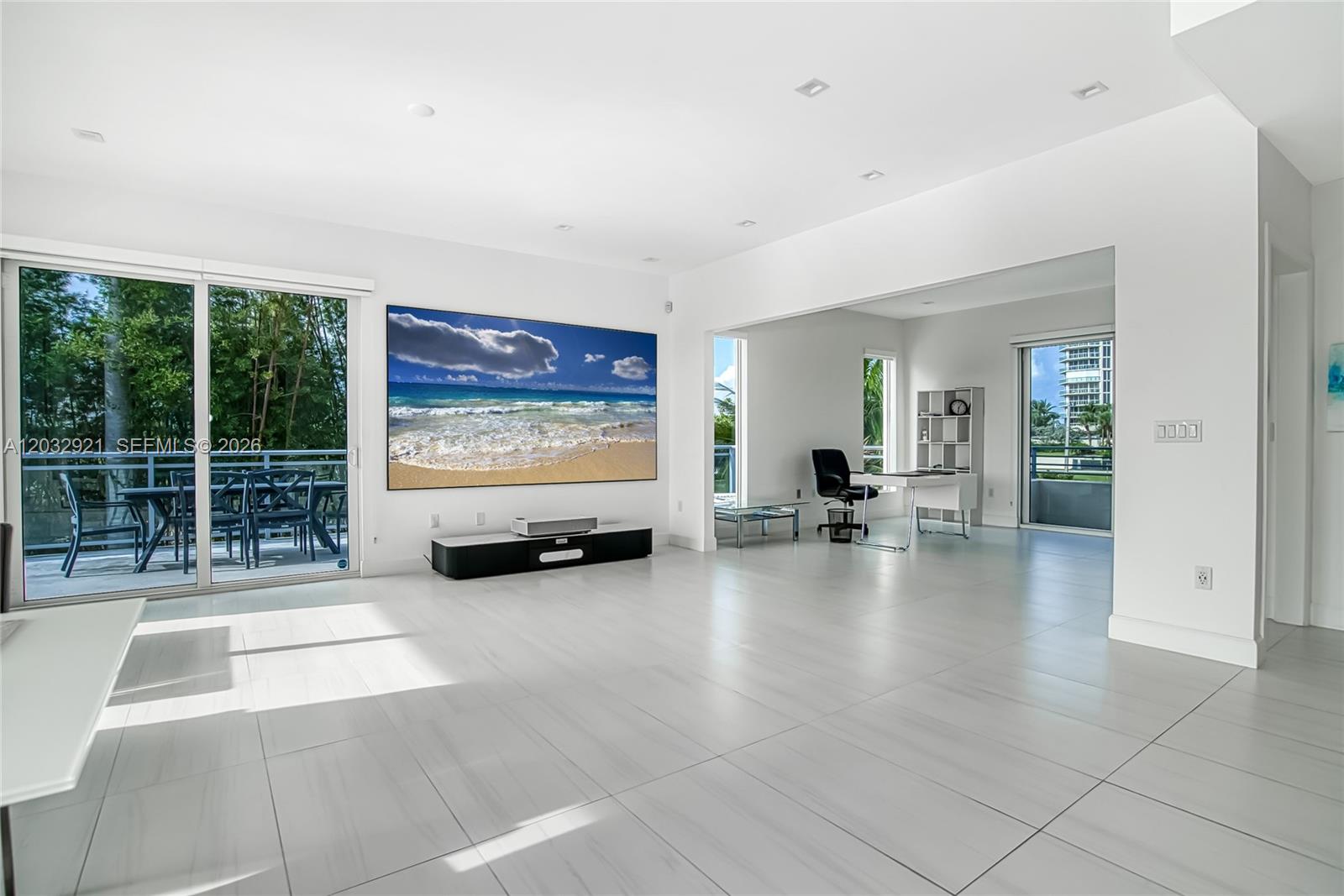 A bright, minimalist living area features white tile floors, a large wall-mounted television, and an adjacent office — HOLLYWOOD, FL