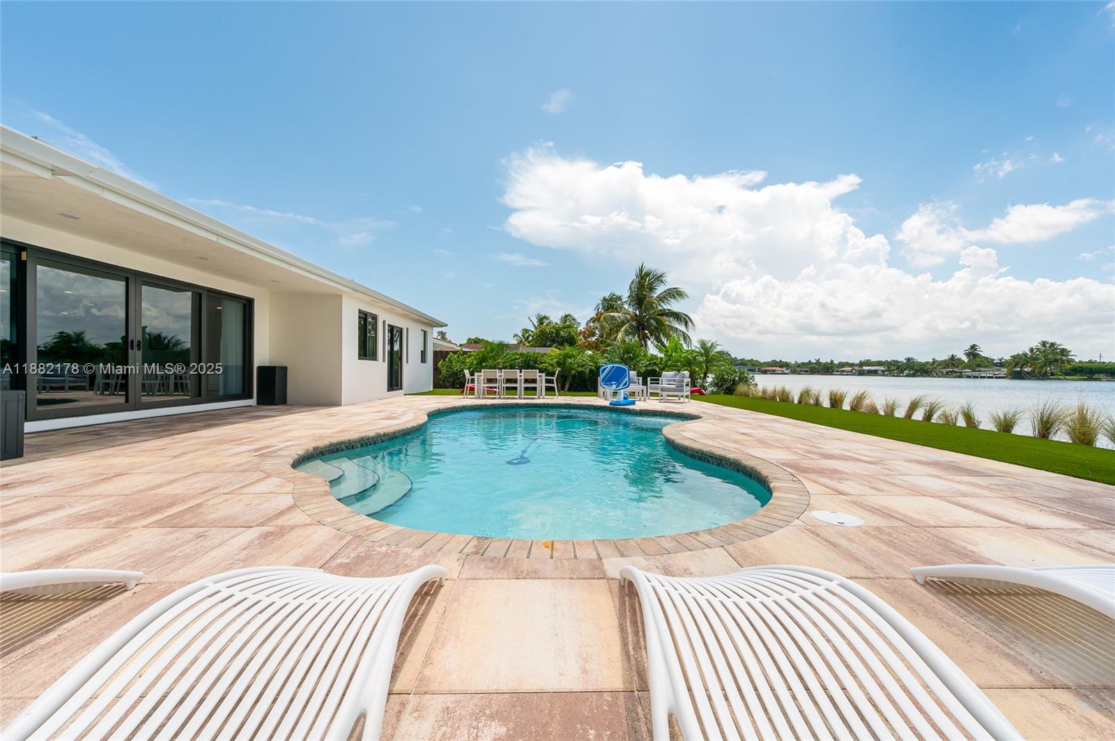 A patio with a swimming pool overlooks a lake under a blue sky — 21230 NE 25th Ct, Miami FL 33180, MIAMI, FL