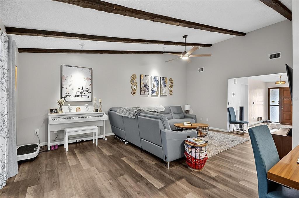 A living room features wood-look flooring, exposed ceiling beams, a gray sectional, and a white piano — 3 BLAKE COURT, PALM COAST, FL