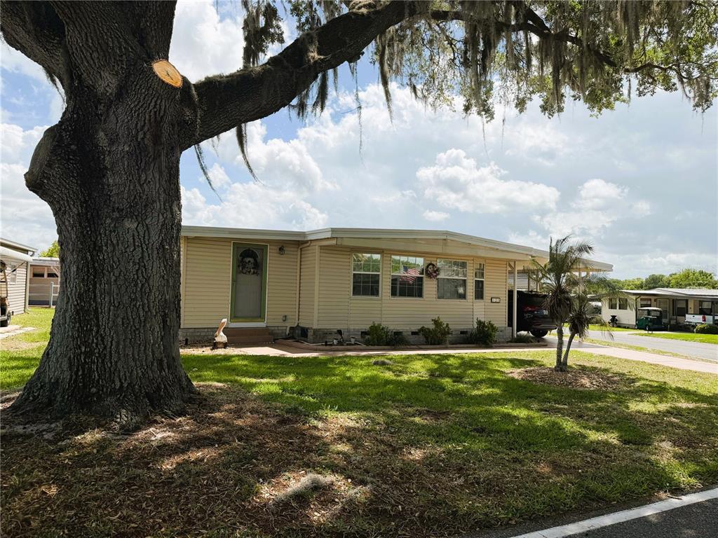 A single-story mobile home with beige siding sits under a large oak tree — 121 AZALEA TRAIL, LEESBURG, FL