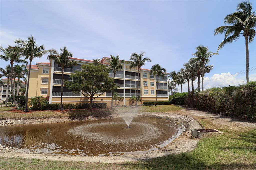A multi-story yellow apartment building stands behind a small pond with a water fountain — 255 W END DRIVE UNIT 2210, PUNTA GORDA, FL