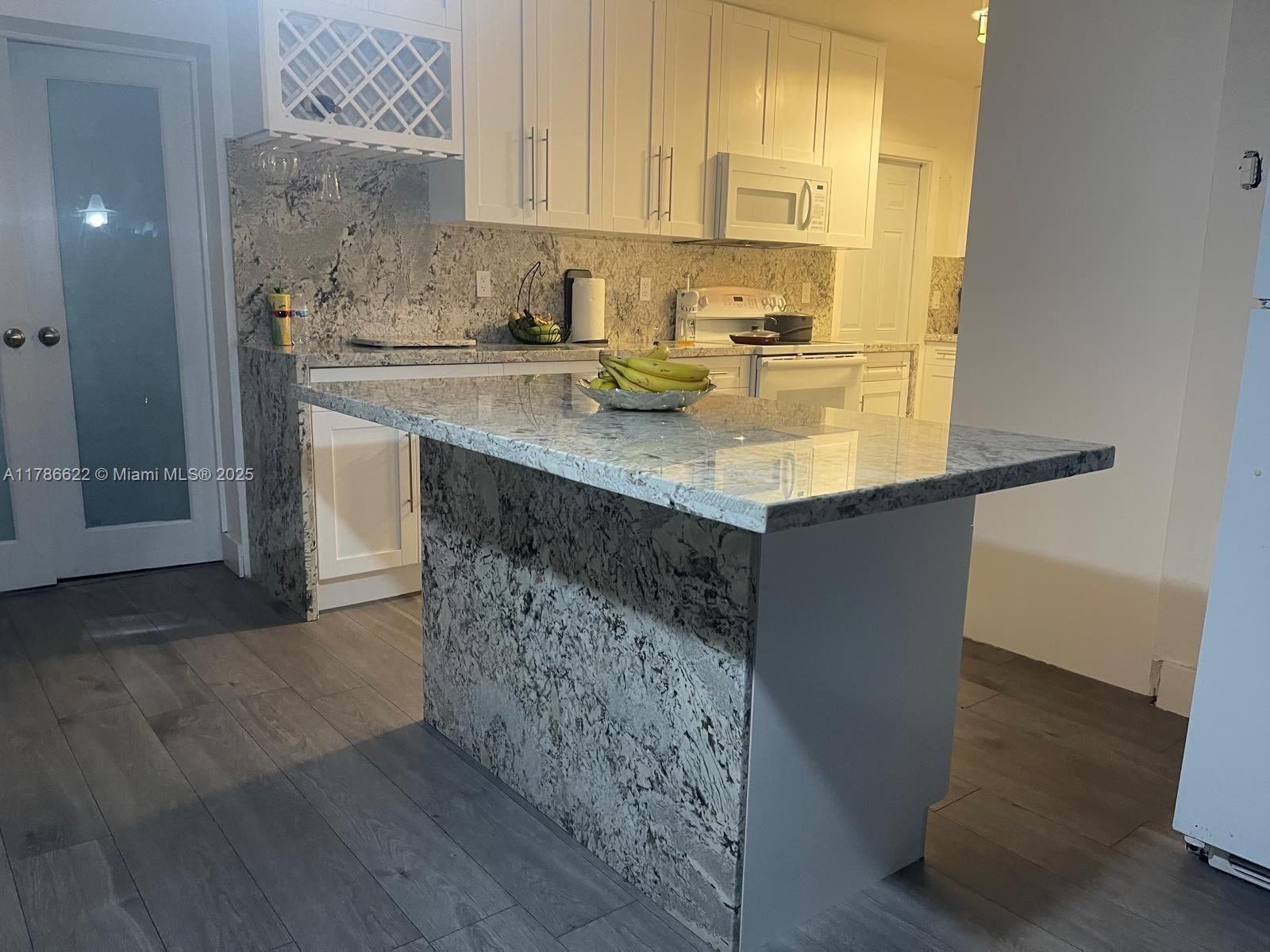 A kitchen features white cabinetry, granite countertops, a central island, and wood-look flooring — 3103 NW 157th St, Miami Gardens FL 33054, MIAMI GARDENS, FL