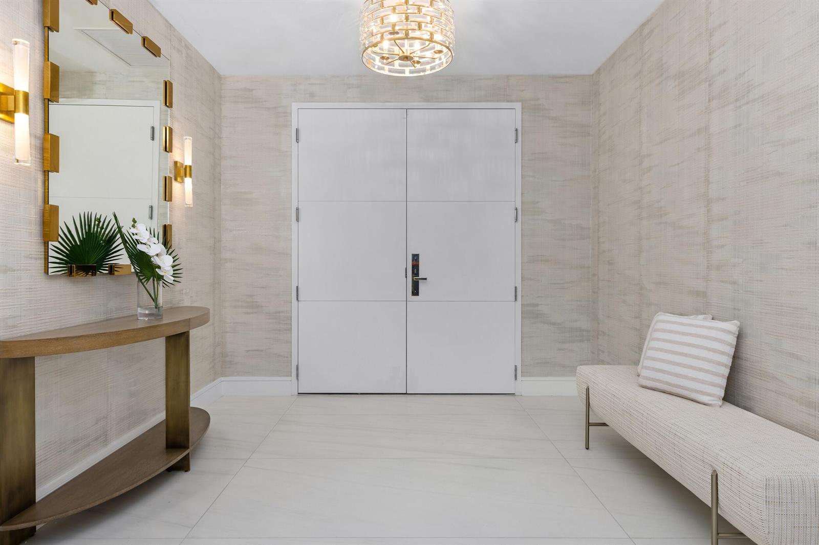 A modern entryway features a white double door, textured wallpaper, a console table, and bench — 725 S Ocean Boulevard # 402, Boca Raton FL 33432, BOCA RATON, FL