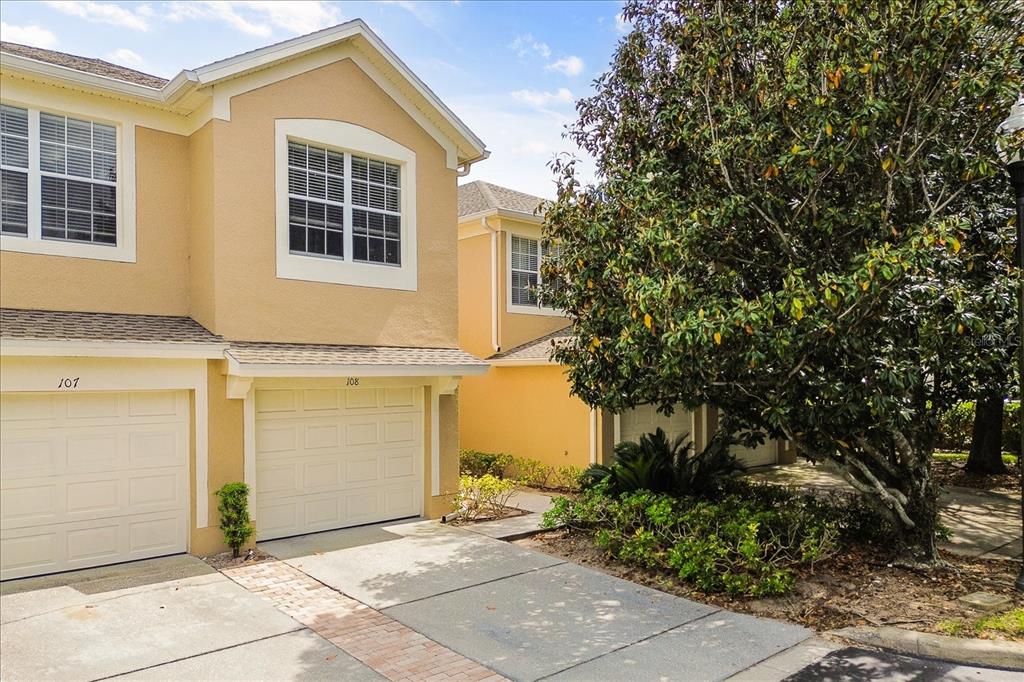 Two tan townhouses with white garage doors and driveways stand next to a large tree — 6440 AXEITOS TERRACE UNIT 108, ORLANDO, FL