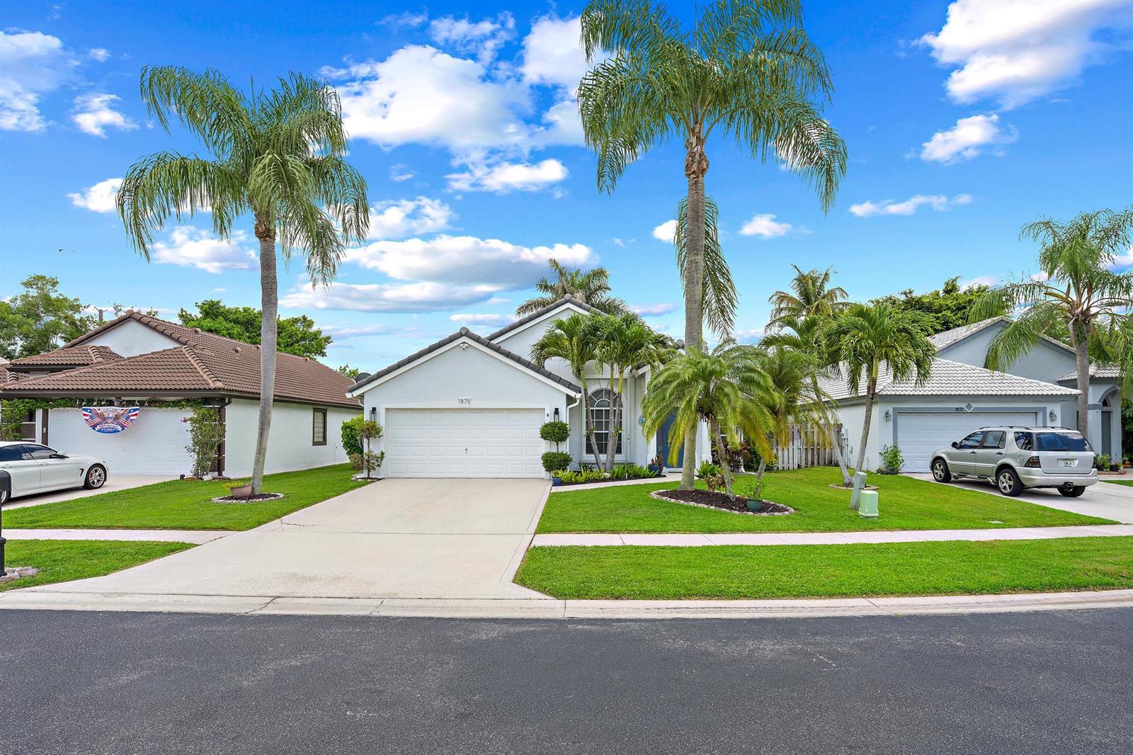 A single-story suburban house with a white garage, palm trees, and a green lawn — 1875 Oak Berry Circle, Wellington FL 33414, WELLINGTON, FL