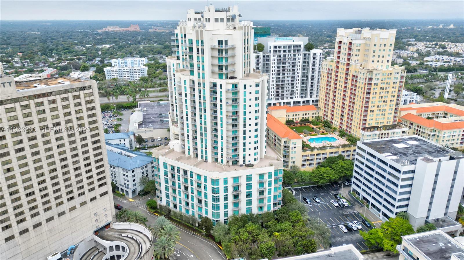 An aerial view shows a tall, white residential skyscraper surrounded by other urban buildings and parking — 9025 S DADELAND BLVD, Miami FL 33156, MIAMI, FL
