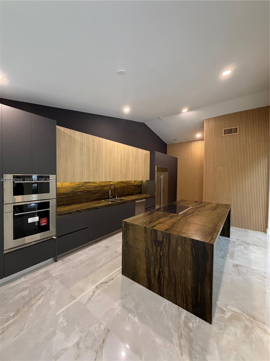 A modern kitchen features dark cabinetry, wood-paneled walls, a stone island, and marble flooring — 4499 NW 93rd Doral Ct, Doral FL 33178, DORAL, FL