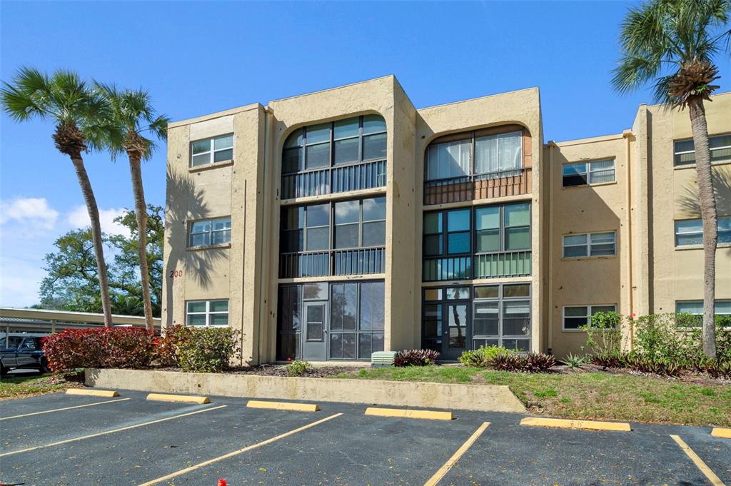 A three-story tan apartment building features screened balconies and a parking lot in front — 11485 OAKHURST ROAD UNIT 200-208, LARGO, FL
