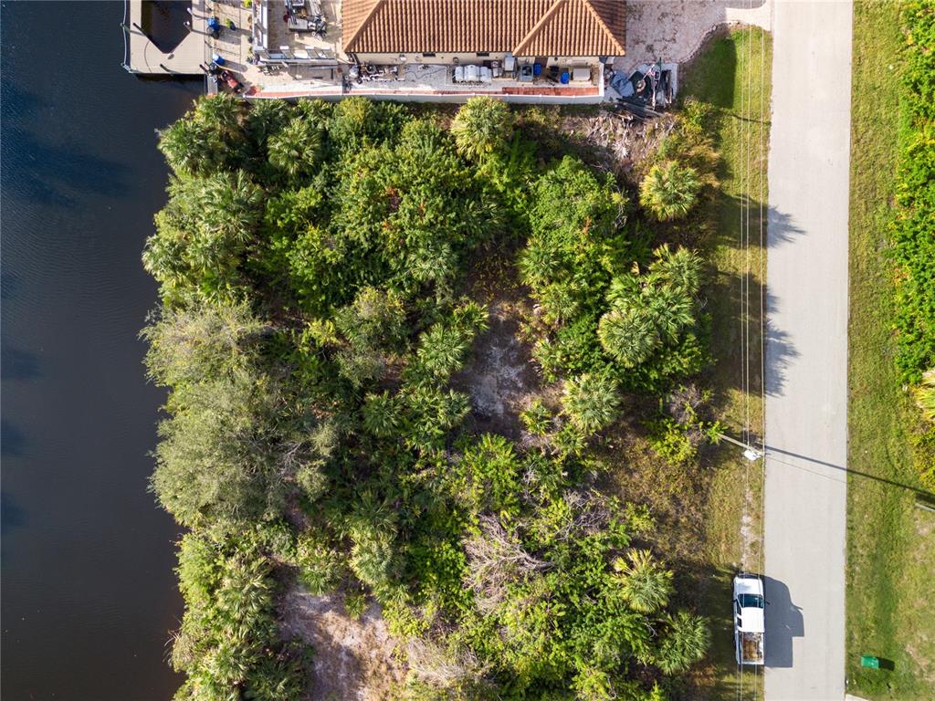 An aerial view shows a vacant, tree-covered waterfront lot next to a house and road — 13886 ALLAMANDA CIRCLE, PORT CHARLOTTE, FL