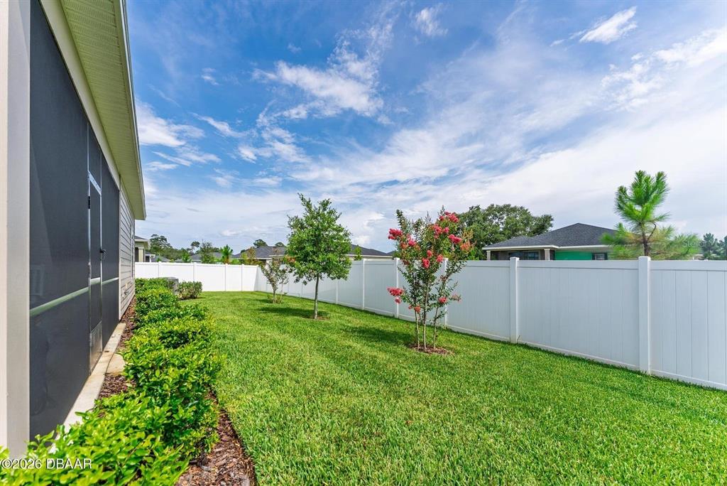 A grassy backyard features a white vinyl fence, small trees, and a screened-in patio — 4116 ACOMA DRIVE, ORMOND BEACH, FL