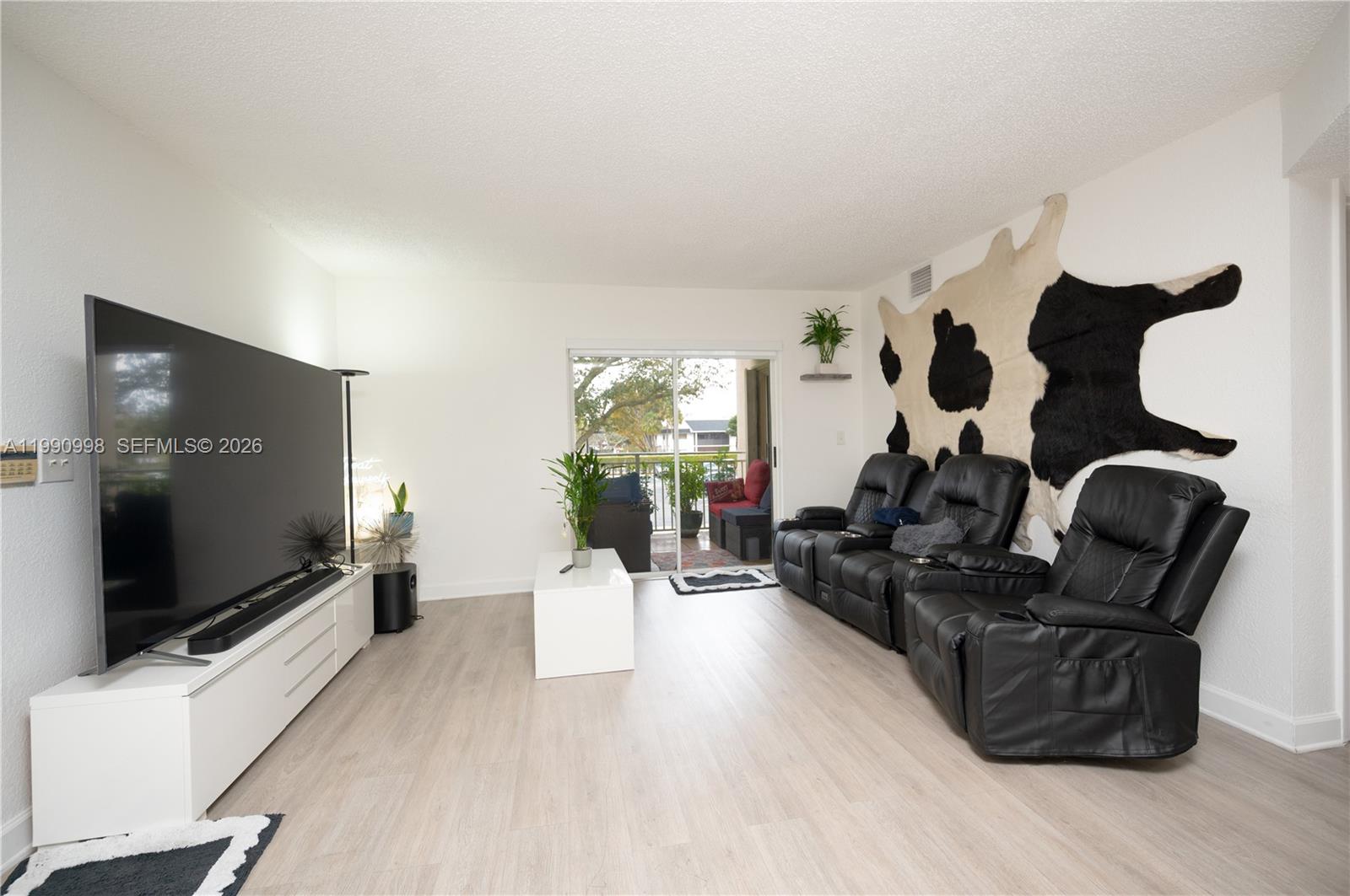 A living room features light wood-look flooring, a large television, and black leather reclining chairs — 18890 NW 57 ave # 210, Hialeah FL 33015, HIALEAH, FL