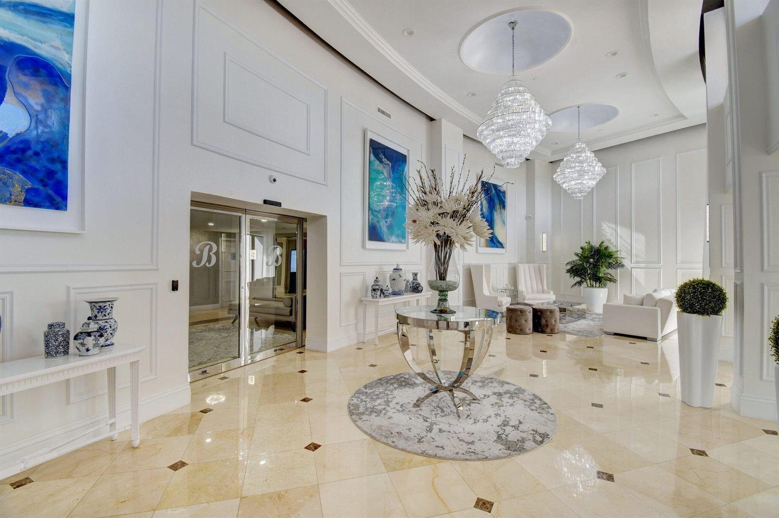 A bright, elegant lobby features marble floors, crystal chandeliers, and white walls with blue artwork — 4740 S Ocean Boulevard # 1503, Boca Raton FL 33487, BOCA RATON, FL