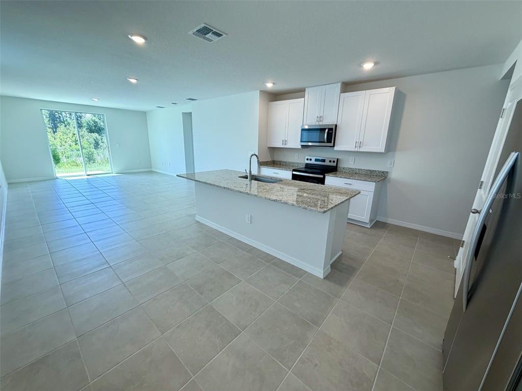 A kitchen with white cabinets, granite countertops, and an island opens to a tiled living area — 1483 BREKEY WAY, ZEPHYRHILLS, FL
