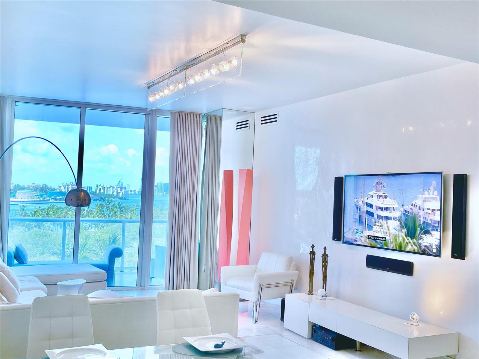 A bright living room features white furniture, a wall-mounted television, and floor-to-ceiling windows with views — 100 S Pointe Drive # 602, Miami Beach FL 33139, MIAMI BEACH, FL