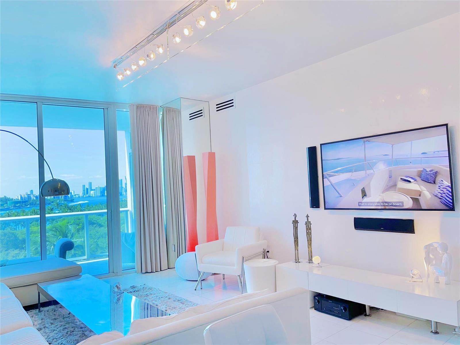 A bright living room features white furniture, a wall-mounted television, and floor-to-ceiling windows with city views — 100 S Pointe Drive # 602, Miami Beach FL 33139, MIAMI BEACH, FL
