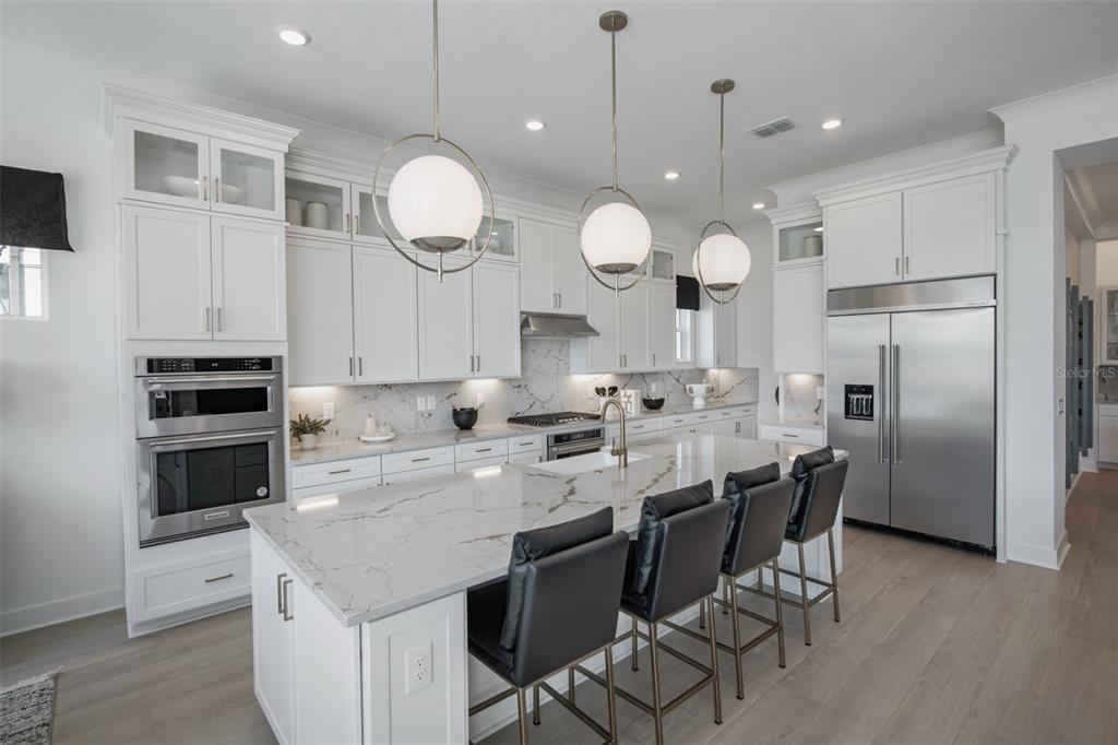 A modern white kitchen features a large marble-topped island with black bar stools and stainless-steel appliances — 35176 COHAGEN TRAIL, ZEPHYRHILLS, FL