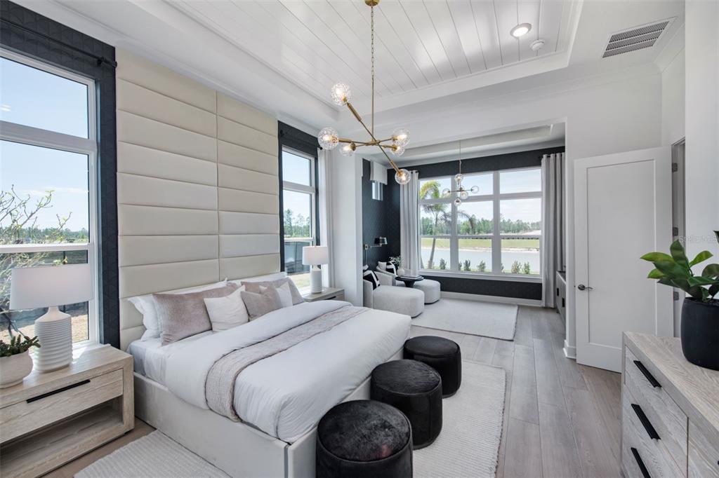 A modern bedroom features a tufted headboard, a chandelier, and large windows overlooking a lake — 35176 COHAGEN TRAIL, ZEPHYRHILLS, FL