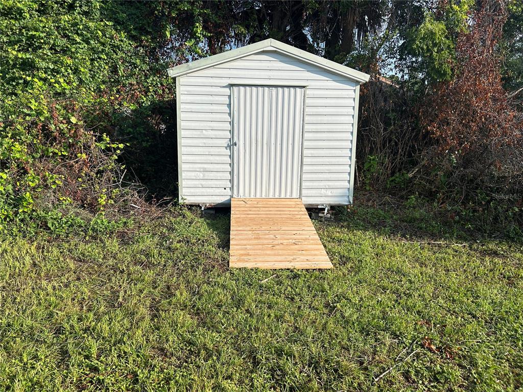 A white metal storage shed with a wooden ramp sits in a grassy yard — 1450 AGATHA DRIVE, DELTONA, FL