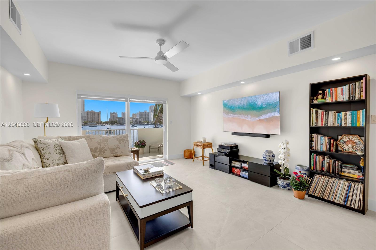A bright living room features a sofa, coffee table, wall-mounted television, and a tall bookshelf — 462 Golden Isles Dr # 211, Hallandale Beach FL 33009, HALLANDALE BEACH, FL