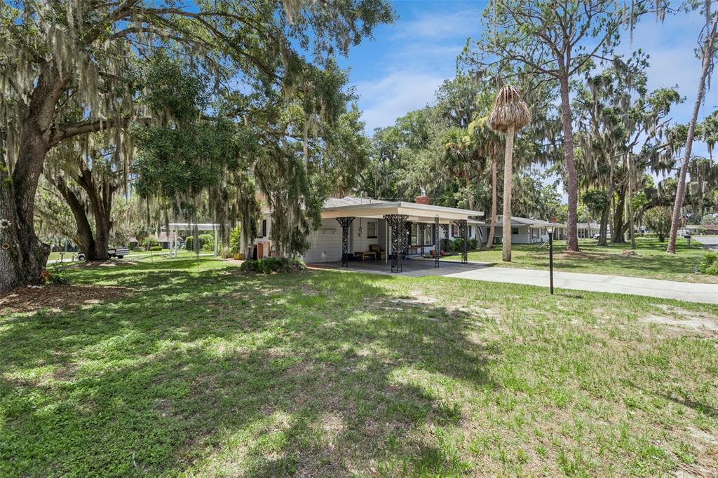 A single-story house with a carport sits on a grassy lot shaded by large trees — 1400 SPARTAN AVENUE, LEESBURG, FL