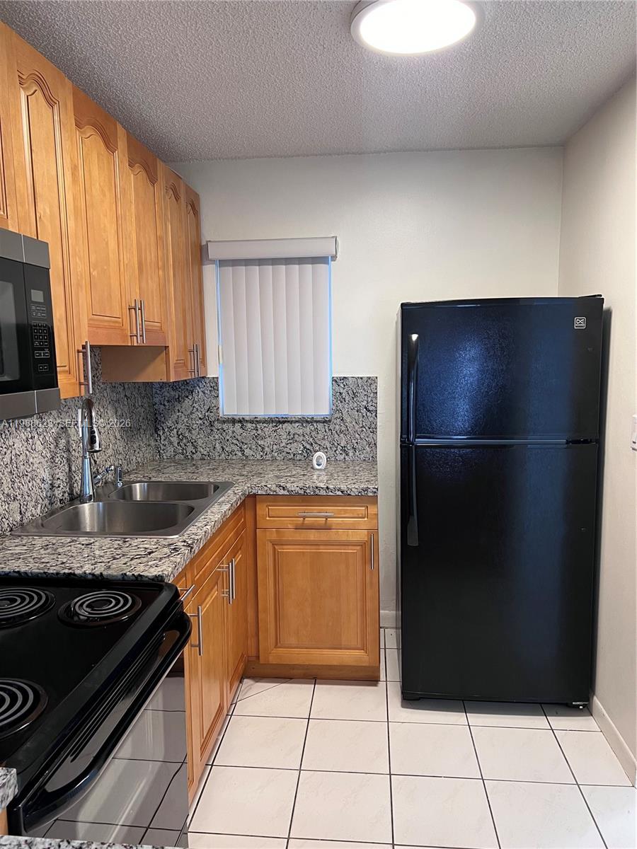 A kitchen features light wood cabinets, granite countertops, a black refrigerator, and white tile flooring — 2750 W 76th St # 208, Hialeah FL 33016, HIALEAH, FL