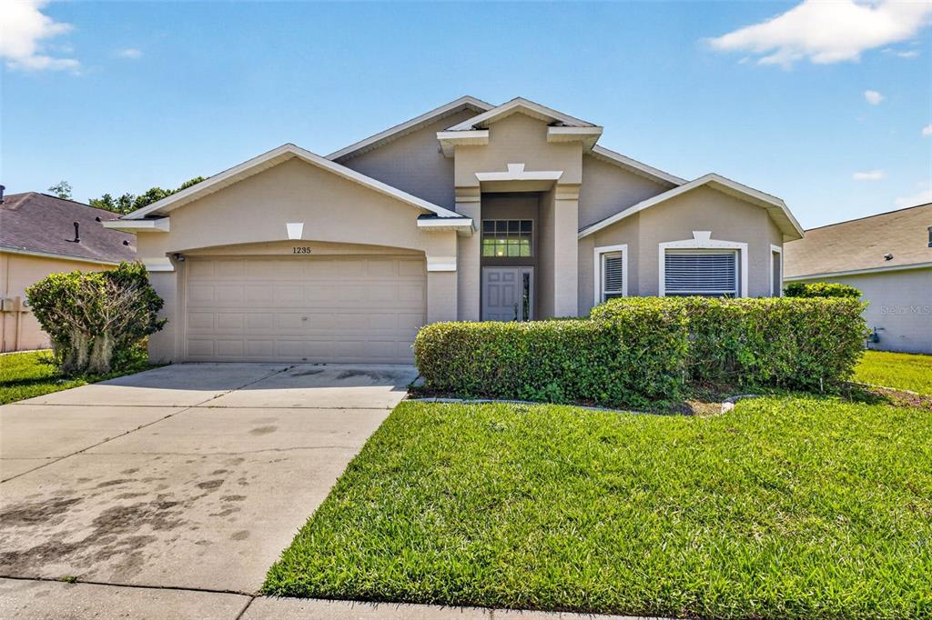 A single-story suburban house with a two-car garage, beige stucco exterior, and front lawn — 1235 KATAHDIN COURT, WESLEY CHAPEL, FL