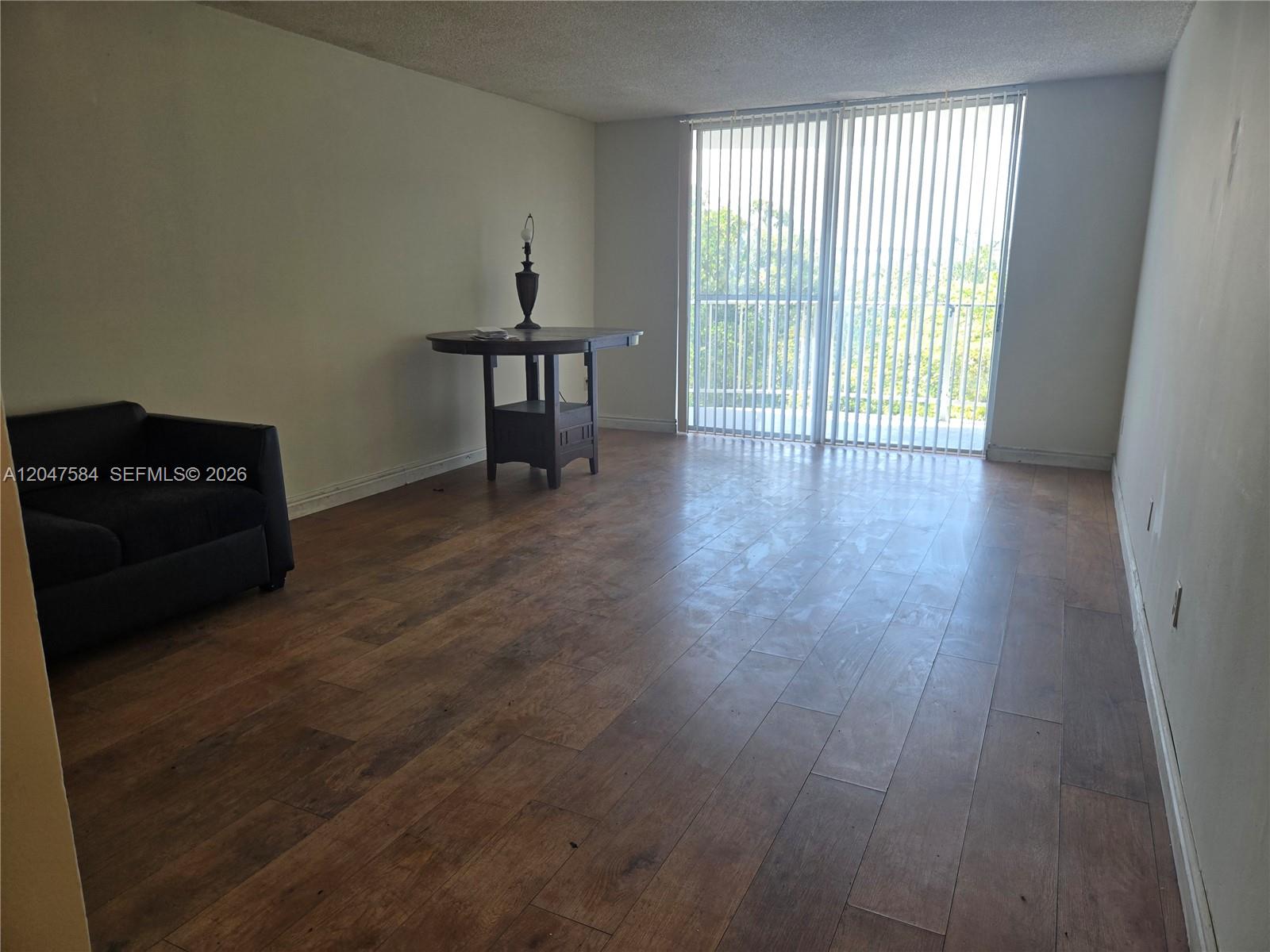 An empty living room features wood-look flooring, a dark armchair, a small table, and vertical blinds — 482 NW 165th St Rd # 505, Miami FL 33169, MIAMI, FL