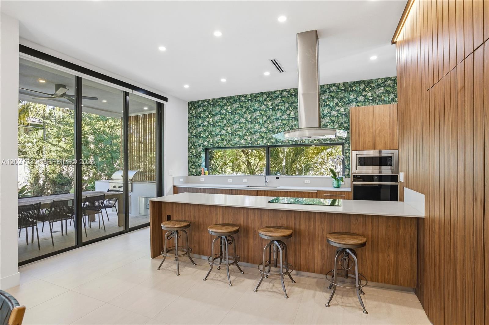 A modern kitchen features wood cabinetry, a white island with barstools, and tropical-patterned wallpaper — 4354 Alton Rd, Miami Beach FL 33140, MIAMI BEACH, FL
