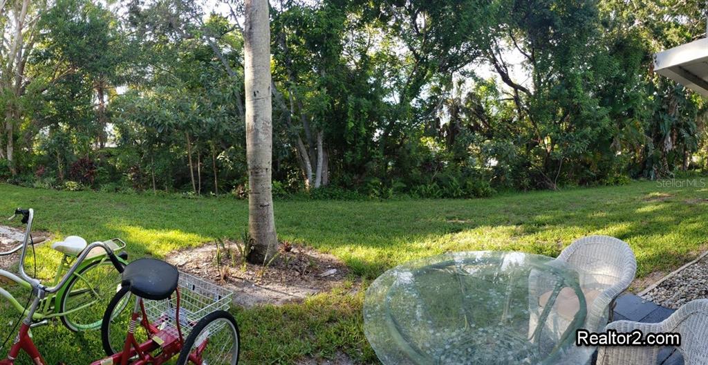 A backyard view features a grassy lawn, dense trees, a bicycle, and patio furniture — 2047 CANAL DRIVE UNIT L6, BRADENTON, FL