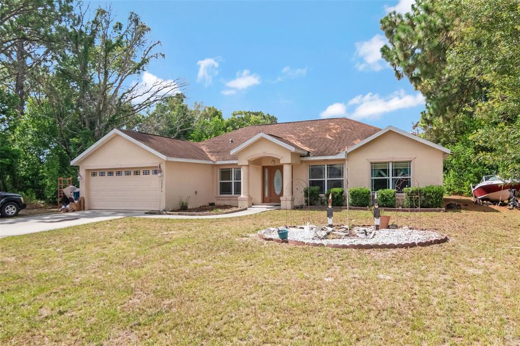 A single-story tan house with a two-car garage and a front yard garden bed — 12276 VILLA ROAD, SPRING HILL, FL